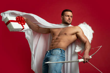 Sexy man on valentines day. Handsome angel man with angel wings. Great present for girlfriend. Charms of love. Valentine gift. Sexy guy on red background. Cupid shoot with bow and arrowsの写真素材