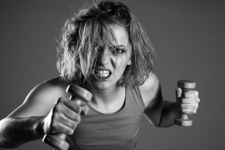 Woman coach with angry face holds dumbbells n grey background. Training hard, overcomes pain. Excited girl does exercise with dumbbells. Woman power concept.の写真素材