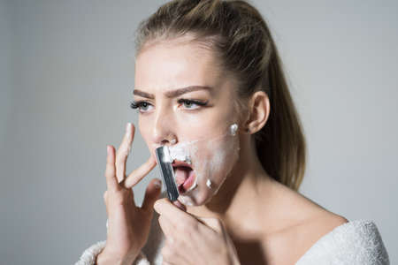 Girl moustache. Barber and shaving concept. Woman with face covered with foam holds straight razor in hand. Girl on busy face wears bathrobe. Lady shaves her face with sharp blade of straight razor.の写真素材