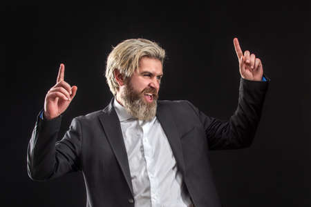 Happy attrative young businessman in black suit listening music with earphones and dancing over gray...