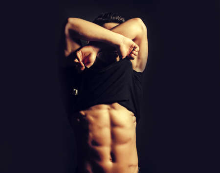 Sexy man in black shirt with fashionable hairdo undressing with bare muscular torso and beautiful body indoor on studio backgroundの写真素材