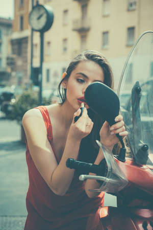 Woman applying makeup on the street. Sexy fashion girl model with red lips put lipstick looking in a mirror motorcycle.の写真素材