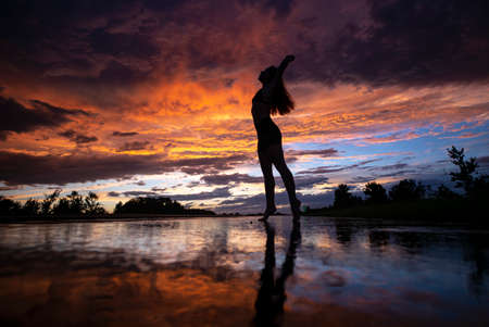 Black silhouette of pretty girl or beautiful woman, sexy slim model, with long hair poses on sea beach at sunset outdoors on violet sky backgroundの写真素材