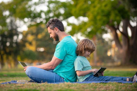 Kid with daddy learning lesson. Father and son use laptop, dad and school boy child looking tablet. Outdoor distance learning.の写真素材