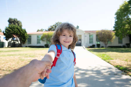 Little boy holding parent fathers hand with backpack on blurred school building background.