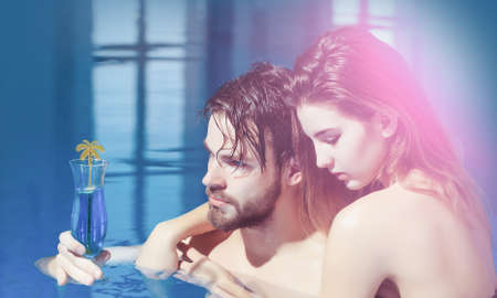 Couple in pool of pretty woman or sexy girl and handsome bearded man or guy with muscular body in swimming pool with blue water holds cocktail glass.の写真素材