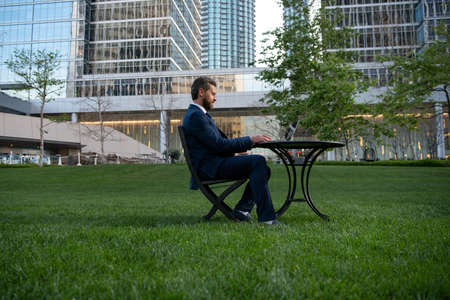 Middle aged businessman typing on laptop computer sitting at table on office terrace. Man working remotely in modern park.の写真素材