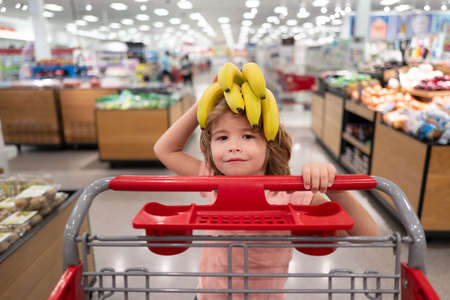 Funny kid with shopping cart buying food at grocery store or supermarket. Surprised kids face. Banana on funny child head.の写真素材