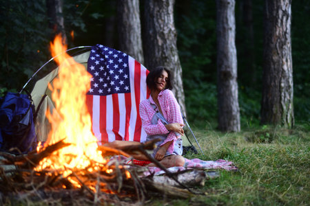 Beautiful dreamy girl enjoy warm bonfire and camping in the forest. Sensual girl sitting near fire. Traveling and camping concept. American flag hang at camping tent.の写真素材