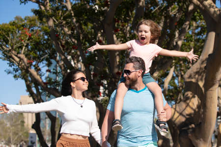 Happy family vacation. Joyful father giving piggyback ride to his son. Happy parents holding hands with son and walking in sunny summer citi street, tender family moment.の写真素材