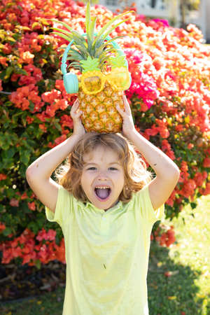 Funny child boy with a pineapple on head in green summer park.