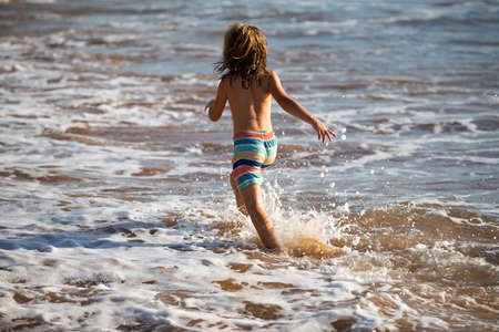Back view of kid boy have fun on tropical sea beach. Funny child run with splashes by water pool...