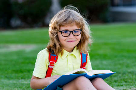 Back to school. Funny little boy in glasses near school. Child from elementary school with book and bag. Education. Child with a book.の写真素材