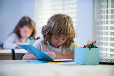 School kid writing something in copybook and sitting at table in classroom. Portrait of child boy...