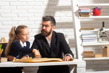 Elementary funny pupil girl with serious teacher in classroom. Fathers and daughters learning. Doing homework with dad.の写真素材