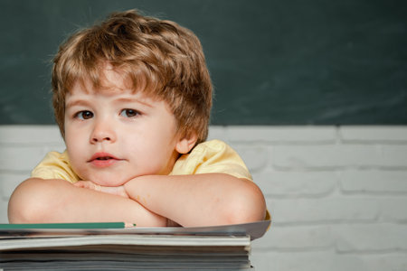 Boy pupil with happy face expression near desk with school supplies. Child is ready to answer with a blackboard on a background. Teachers day.の写真素材