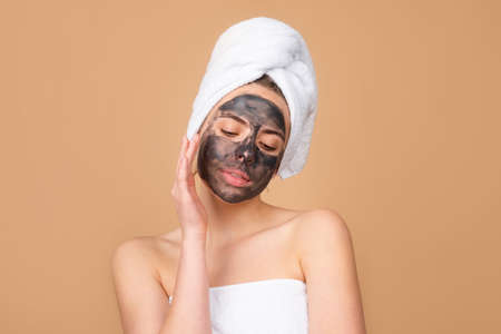 Woman with with charcoal mask touching her nose. Mud facial mask, face clay mask spa. Beautiful woman with cosmetic mud facial procedure, spa health concept. Skin care beauty treatment. Towel on head.の写真素材