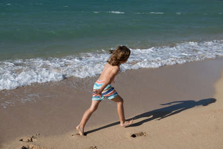 Happy kid boy have fun on tropical sea beach. Funny child run with splashes by water pool along surf edge. Kids activity on summer holiday.の写真素材