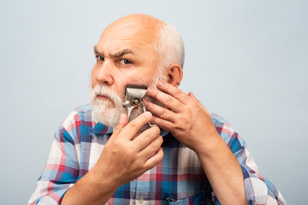 Old gray man cut bear with hair clipper. Portrait of barber hold old razor hair clipper, vintage barber shop, shaving. Old bearded man. Vintage barbershop.の写真素材