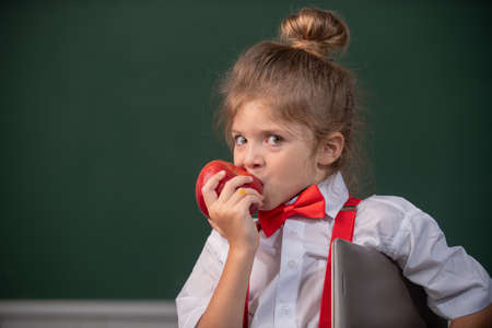 Little schoolgirl pupil student in class, eating apple at school. Funny kids face close up.の写真素材
