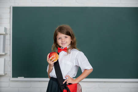 Happy smiling preteen girl, kid schoolgirl with wears schhol uniform holding backpack on blackboard background looking at camera. Education, learning and children conceptの写真素材