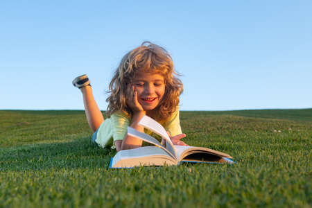 Funny kid reading the book in spring park. Child boy reading book, laying on grass in field on sky background. Portrait of clever kids.の写真素材