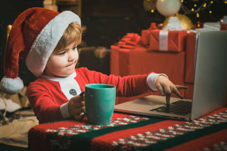Online Christmas shopping for kids. Little child boy is wearing Santa clothes sitting by his laptop....
