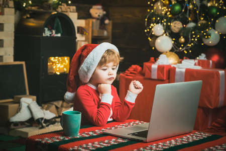 Christmas child boy with computer laptop at home on Christmas background. Little kid is wearing Santa clothes sitting by his laptop. Santa helper using notebook.の写真素材