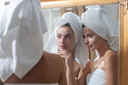 Beautiful woman look in mirror after shower in apartment or hotel room. Young pretty girls friends wrapped in towel smoothing perfecting skin, daily morning routine concept.の写真素材