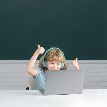 Smart boy with thumbs up in headphones sit at desk, study online on laptop at school, klever kid wear earphones in notebook learning using internet lessons. First day at school.の写真素材