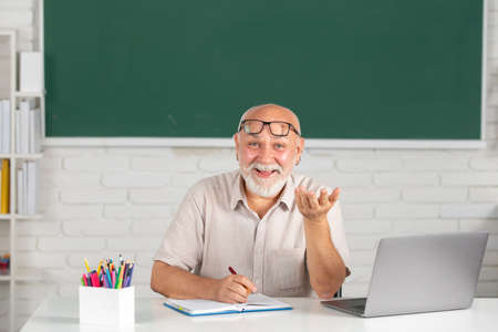 Man mature teacher. Portrait of happy senior tutor in glasses with book on the blackboard in class at high school or collage. Old student study in university. Adult teaching.の写真素材