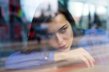Romantic casual woman portrait. Thoughtful concept. Woman at a cafe while gazing through the window glass. Sad alone girl. Beautiful girl in the window watching.の写真素材