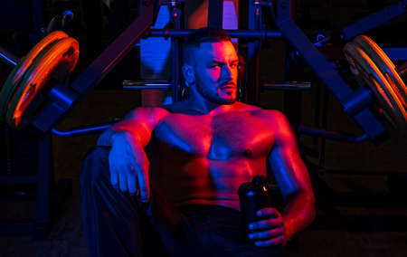 Fit man. Young muscular man with naked torso working out in gym. Resting time. Tired sporty man resting after training. Man with bottle of water or protein.の写真素材