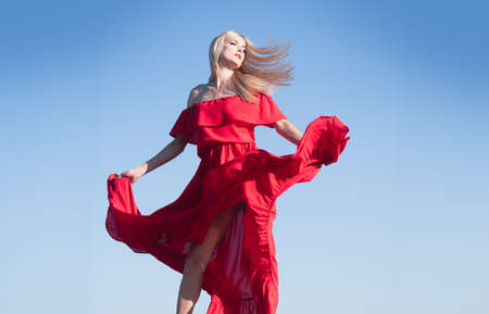 Blonde woman in red dress movement on sky. Fashionable young model in style dress. Fashion woman in summer.の写真素材