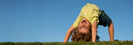 Banner with spring kids portrait. Excited little boy laying upside on the grass in the park. Outdoor portrait of pretty little boy on fresh green grass. Active healthy outdoor sport. Fun activity.の写真素材