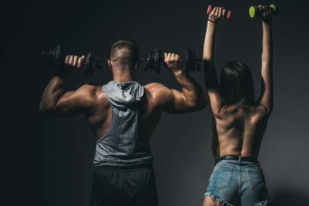 Sexy sport couple exercising with dumbbell. Muscular man with naked body, fitness woman with dumbbells on a dark background, back view. Slim sexy girl with strong muscular man workout.の写真素材