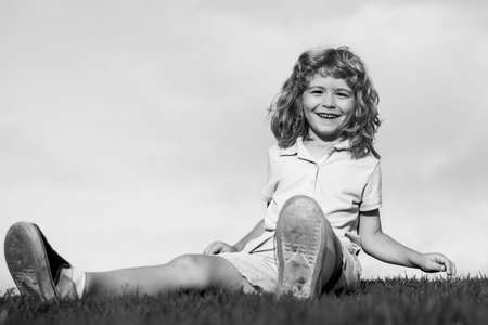Happy child boy sitting on green grass outdoors in summer park.