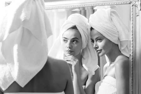 Beautiful woman look in mirror after shower in apartment or hotel room. Young pretty girls friends wrapped in towel smoothing perfecting skin, daily morning routine concept.の写真素材