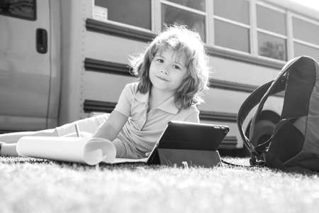 Smart school boy doing homework with digital tablet at school park near school bus. Child using...