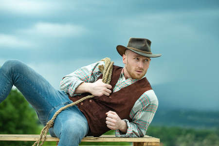 Cowboy on ranch. Handsome man in cowboy hat and retro vintage outfit. Cowboy with lasso rope on sky background.の写真素材