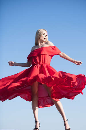 Blonde woman in red dress movement on sky. Attractive woman in fashion outfit outside. Outdoor fashion photo of young beautiful lady enjoying spring. Carefree women outside.の写真素材