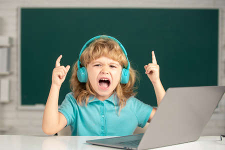Angry boy in headphones sit at desk, study online on laptop at school, klever kid wear earphones in...