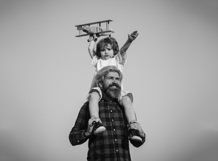 Fathers day. Father and son. Father giving son ride on back. Portrait of happy father giving son piggyback ride on his shoulders and looking up. Cute boy with dad playing with wooden airplane outdoor.の写真素材