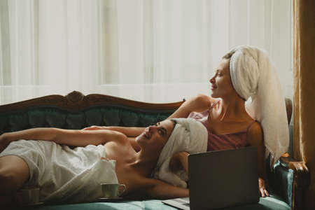 Relaxed young female friends with coffee cup and laptop in bed at home. Cheerful sexy girl with bathrobes towels on head in bedroom after morning wake up.の写真素材