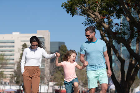 Happy parents holding hands with son and walking in sunny summer citi street, tender family moment. Family in urban skyline on park lawn.の写真素材