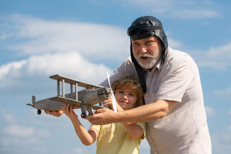 Old grandfather and young child grandson playing with wooden plane against summer sky background. Child boy with dreams of flying or traveling.の写真素材