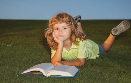 School boy with a book having a rest outdoor. Clever child boy reading book laying on grass on grass and sky background with copy space.の写真素材