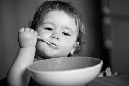 Portrait of funny little baby boy eating from plate holding spoon closeup. Baby holding a spoon while putting eat in his mouth.の写真素材