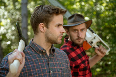 Lumberjack workers in the forest. Woodcutter with axe and lumberjack with chainsaw. Handsome woodcutter men hipsters in forestの写真素材