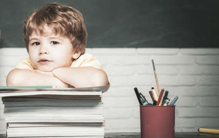 Little boy pupil with happy face expression near desk with school supplies. Child is ready to answer with a blackboard on a background. Teachers day.の写真素材